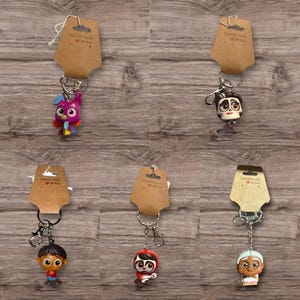 May include: Keychains featuring colorful character figurines, each attached to a metal keyring and a brown paper tag that reads "Handmade with love". The keychains are displayed on a wooden surface.