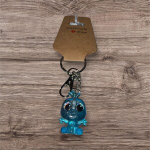 May include: A blue keyring with a small, glittery figure with large eyes and a smiling face. The keyring includes a metal clasp, a chain, and a tag that reads "Handmade with love."