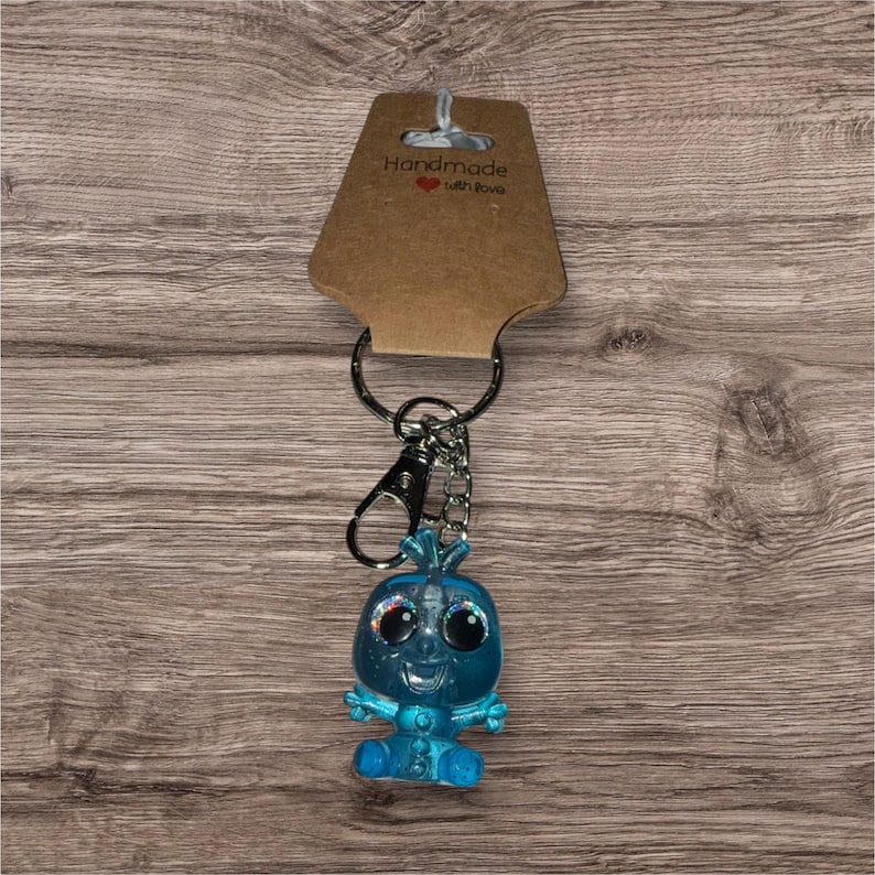 May include: A blue keyring with a small, glittery figure with large eyes and a smiling face. The keyring includes a metal clasp, a chain, and a tag that reads "Handmade with love."