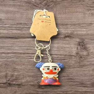 Pu&ograve; includere: Un portachiavi a forma di clown con catena e chiusura argentate. Il clown ha un viso bianco, capelli blu, naso e bocca rossi e indossa una camicia viola e scarpe arancioni. Un'etichetta marrone con la scritta "Handmade with love" &egrave; attaccata.