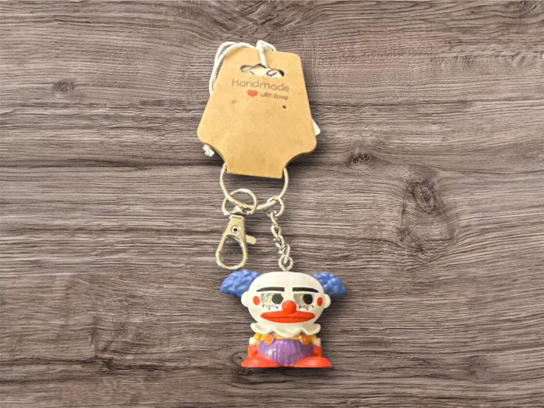 Pu&ograve; includere: Un portachiavi a forma di clown con catena e chiusura argentate. Il clown ha un viso bianco, capelli blu, naso e bocca rossi e indossa una camicia viola e scarpe arancioni. Un'etichetta marrone con la scritta "Handmade with love" &egrave; attaccata.