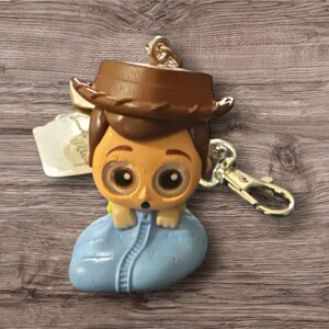 May include: A keychain featuring a cartoon character with a brown cowboy hat, tan skin, and large eyes. The character is wearing a blue outfit and is attached to a silver-colored clasp.