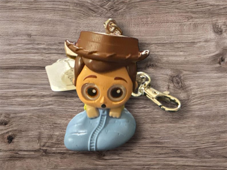 May include: A keychain featuring a cartoon character with a brown cowboy hat, tan skin, and large eyes. The character is wearing a blue outfit and is attached to a silver-colored clasp.