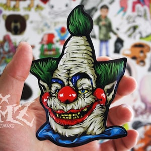 May include: A sticker of a creepy clown with green hair, a blue collar, and a large red nose. The clown has a wide, toothy grin and a sinister expression.