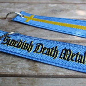 May include: Two blue embroidered keychains. One features the Swedish flag with a yellow cross. The other keychain has the text "Swedish Death Metal" in black and yellow lettering. Both keychains have a silver key ring.