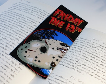May include: A red and black bookmark featuring a white hockey mask and the text "FRIDAY THE 13th" in red and yellow lettering. The background is a blue sky with a pine tree.