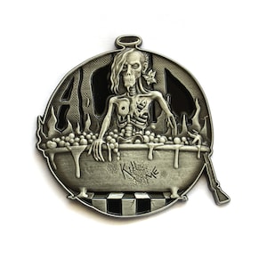 Pode incluir: Um alfinete de metal prateado com um esqueleto numa banheira com o texto "Kill Me". O alfinete tem um halo acima da cabeça do esqueleto e um rifle na lateral. O fundo é preto.