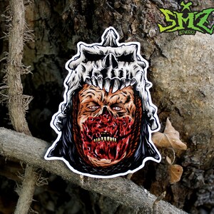 Könnte beinhalten: Ein Aufkleber mit einer detaillierten Illustration einer monströsen, blutigen und skelettartigen Kreatur mit einem Schädelhelm. Die Kreatur hat ein rotes, blutiges Gesicht mit scharfen Zähnen und einem bedrohlichen Ausdruck. Der Aufkleber befindet sich auf einem braunen, strukturierten Hintergrund.