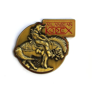 Könnte beinhalten: Goldfarbene Metallnadel mit einem Reiter auf einem buckelnden Pferd. Die Nadel hat ein kreisförmiges Design mit einem rechteckigen Oberteil, auf dem in roten Buchstaben "ATLANICAN KODEX" steht. Die Nadel ist wahrscheinlich ein Sammlerstück oder ein Souvenir.