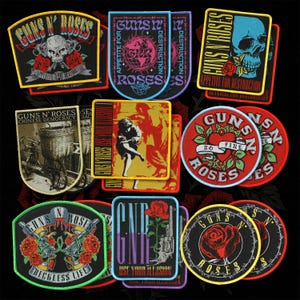 Può includere: Una collezione di toppe ricamate dei Guns N' Roses con vari design di copertine di album, inclusi teschi, rose, chitarre e loghi della band. Le toppe sono circolari, rettangolari e a forma di scudo con colori vivaci rossi, gialli, blu e neri.
