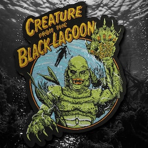 May include: An embroidered patch featuring the Creature from the Black Lagoon in green with orange lips, set against a blue circular background. The text CREATURE FROM THE BLACK LAGOON is arched in yellow above the figure, with a diver silhouette.