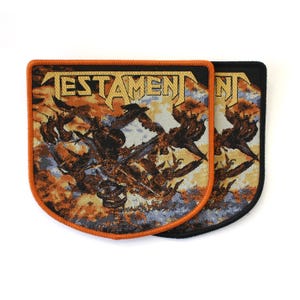 May include: Two rectangular fabric patches with rounded bottom corners featuring the word TESTAMENT in gold lettering. The design displays a dark, chaotic scene with orange, brown, and blue tones. One patch has an orange border, the other black.