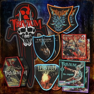 May include: A collection of Trivium band patches featuring various shapes and designs, including a large skull emblem, shield-shaped patches with the text What The Dead Men Say and Ember To Inferno, and rectangular patches with In The Court Of The Dragon.