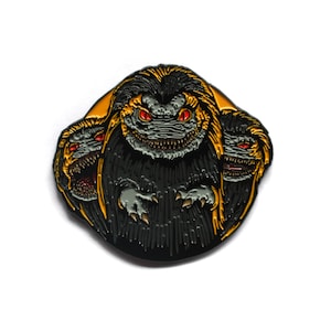 Op de afbeelding: Zwarte emaille pin met een driekoppig monster met rode ogen en scherpe tanden. Het monster wordt omringd door een gele halo.