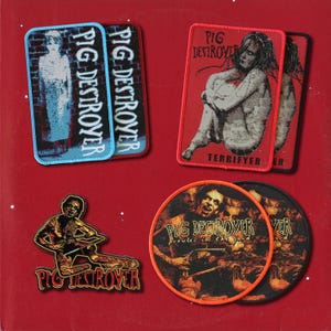 May include: A collection of embroidered patches featuring Pig Destroyer band artwork. Includes two rectangular patches with blue and red borders, a red-bordered rectangular patch, a die-cut figure patch, and two circular patches with orange and black borders.