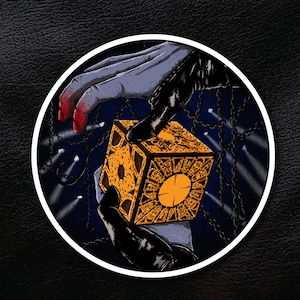 Op de afbeelding: Een hand reikt naar een gouden en zwarte kubuspuzzel met ingewikkelde details. De kubus is omringd door kettingen en een donkere achtergrond.