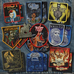 May include: A collection of rectangular, shield-shaped, and circular embroidered fabric patches featuring various band logos and graphic illustrations, including Riot, Cloven Hoof, Accept, Enforcer, Riot City, Sortilège, Sacred Steel, and Mercyful Fate.