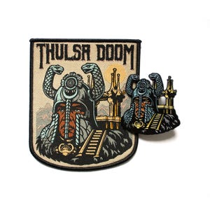 May include: A beige patch and pin featuring the text "THULSA DOOM" above an illustration of a warrior with a serpent-adorned helmet. The warrior has a long beard and is set against a backdrop of mountains and a sun.