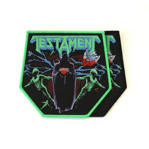 May include: A pentagonal embroidered patch with a bright green stitched border. The design features the word TESTAMENT in light blue, a central dark hooded figure holding a red heart, and two green human-like silhouettes on a black background.