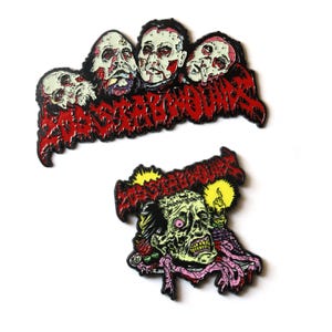 May include: Two enamel pins with a horror theme. One pin features a group of zombie heads with the word "MASSACREBOUND" in red. The second pin shows a zombie head with a candle and the same text. The pins have black outlines.