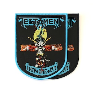 May include: A rectangular embroidered patch with rounded bottom corners, featuring a black background and a light blue border. It displays the word TESTAMENT in light blue, a skull graphic, and the text INTO THE PIT in light blue.