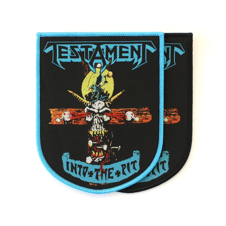 May include: A rectangular embroidered patch with rounded bottom corners, featuring a black background and a light blue border. It displays the word TESTAMENT in light blue, a skull graphic, and the text INTO THE PIT in light blue.