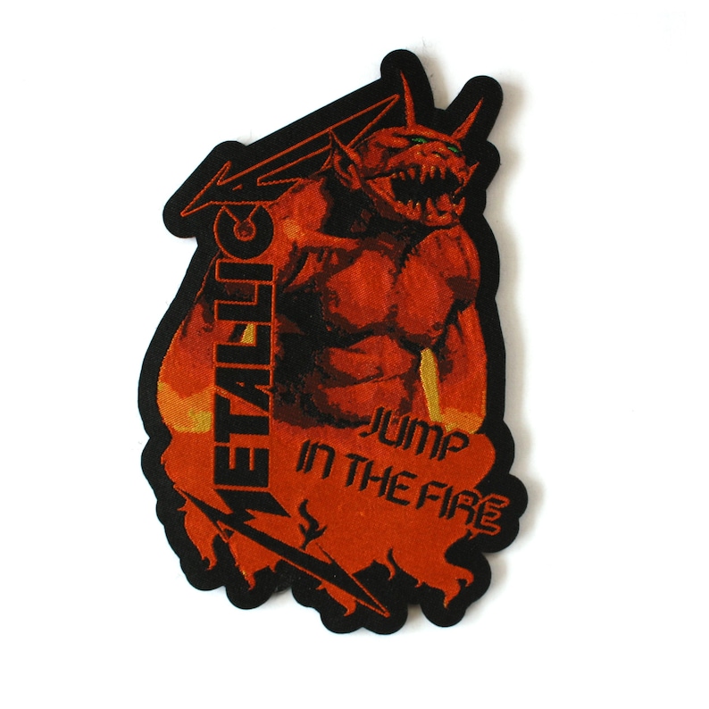 May include: A rectangular embroidered patch with a black border featuring a red and orange demon figure. The text METALLICA is vertically oriented on the left side, and the text JUMP IN THE FIRE is printed in black on the lower right section.
