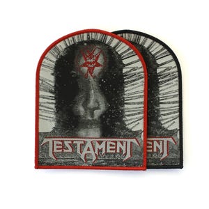 May include: Two arched embroidered patches featuring a black and white graphic of a hooded figure with a red pentagram symbol on the forehead. The word TESTAMENT is printed in a stylized font at the bottom. One patch has a red border, one black.