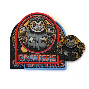 Peut inclure: Trois écussons brodés représentant la créature du film Critters avec des yeux rouges et des dents pointues. Les motifs incluent une bordure circulaire, un fond spatial avec des planètes et une grille. Le texte CRITTERS est brodé en orange.