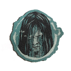 Puede incluir: Un parche circular azul verdoso con un borde blanco bordado. El centro del parche presenta una ilustración en blanco y negro de una mujer con el pelo largo y negro que le cubre la cara. El texto "the ring" está bordado en blanco en el parche.