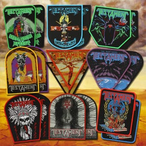 May include: A collection of embroidered fabric patches featuring various Testament band album artwork and logos. The patches are shaped as shields, triangles, and rectangles with colorful graphic designs, including skulls, figures, and text.