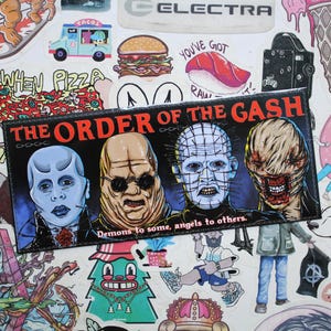 Könnte beinhalten: Ein schwarzer und roter Aufkleber mit dem Text "The Order of the Gash" und einer Grafik von vier Horrorfilmfiguren. Der Text unter der Grafik lautet "Demons to some, angels to others."