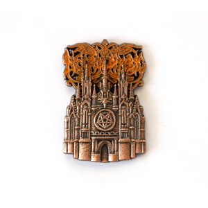 Può includere: Uno spillo in metallo color bronzo con un'illustrazione dettagliata di una cattedrale gotica con un pentagramma al centro. La parte superiore dello spillo presenta il nome della band "NIGHTSHADE" in un font stilizzato con smalto arancione.