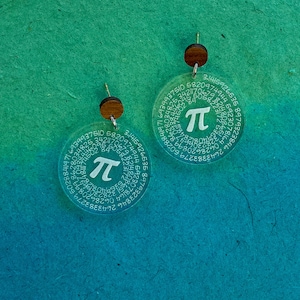 May include: A pair of clear acrylic earrings with a wood backing. The earrings are shaped like circles and have the mathematical constant pi printed on them, along with the first several digits of pi.