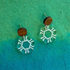 May include: A pair of earrings with a hexagonal design. The earrings are made of clear acrylic with a white geometric pattern. The top of each earring is a brown wooden disc.