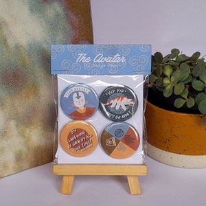 May include: A clear plastic package containing four round Avatar-themed badges. The badges feature cartoon characters and phrases like "Team Avatar" and "Yip Yip, Let's Go Appa!" The package is displayed on a small wooden easel.