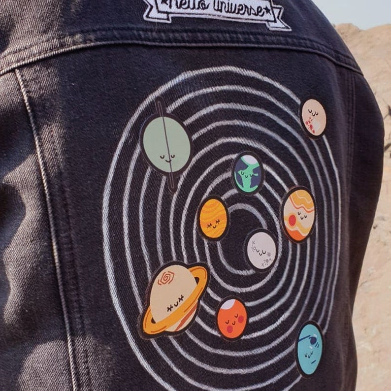 May include: Black denim jacket with a solar system design. The back of the jacket features a white spiral with cartoon planets and the text "hello universe" on a patch at the top. The planets are in various colors.