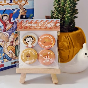 May include: A pack of four round badges featuring characters and phrases from the anime series "One Piece." The badges are in shades of orange, yellow, and white, with text that reads "Monkey D. Luffy," "I'm gonna be king of the pirates," and "Easily distracted by food!"