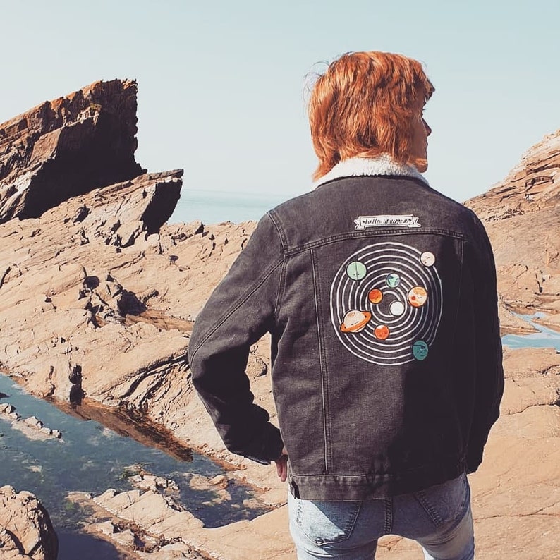 May include: A black denim jacket with a white sherpa collar and a colorful solar system design on the back. The jacket features a small rectangular patch above the solar system graphic with the text "Stella Severa".