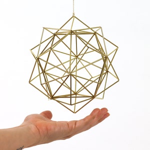 Può includere: Un mobile geometrico dorato con un design complesso e intricato appeso a una catena. Il mobile è sospeso sopra una mano, con il palmo rivolto verso l'alto, come se fosse pronto a prenderlo.