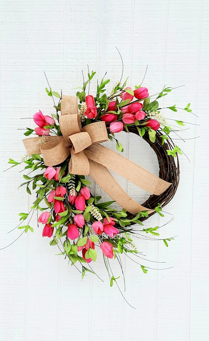 K&ouml;nnte beinhalten: Ein Kranz aus rosa Tulpen mit einer Schleife aus Sackleinen. Der Kranz ist aus Weinreben gefertigt und mit rosa Tulpen und gr&uuml;nen Bl&auml;ttern verziert. Der Kranz h&auml;ngt an einer wei&szlig;en Holzt&uuml;r.