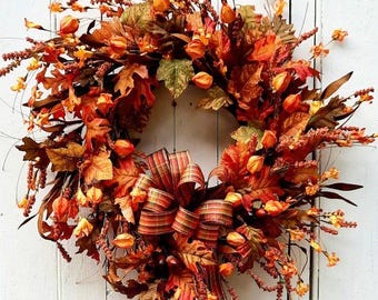 May include: A fall-themed wreath with a variety of autumn leaves in shades of orange, red, and brown. The wreath features a large, multi-layered bow with striped ribbon. Small orange berry clusters and other decorative elements complete the design. The website address "leosgardenhome.com" is visible.