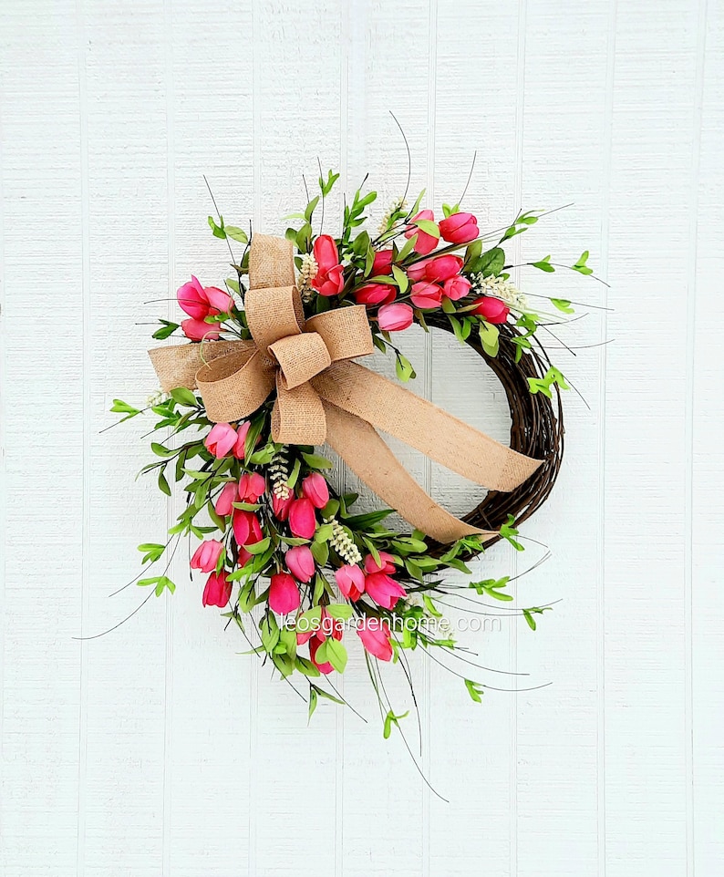 K&ouml;nnte beinhalten: Ein Kranz aus rosa Tulpen mit einer Schleife aus Sackleinen. Der Kranz ist aus Weinreben gefertigt und mit rosa Tulpen und gr&uuml;nen Bl&auml;ttern verziert. Der Kranz h&auml;ngt an einer wei&szlig;en Holzt&uuml;r.