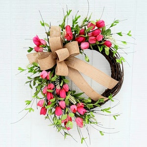 K&ouml;nnte beinhalten: Ein rosafarbener und gr&uuml;ner Blumenkranz mit einer Schleife aus Sackleinen. Der Kranz ist aus Weinreben gefertigt und mit rosa Tulpen und gr&uuml;nen Bl&auml;ttern verziert.