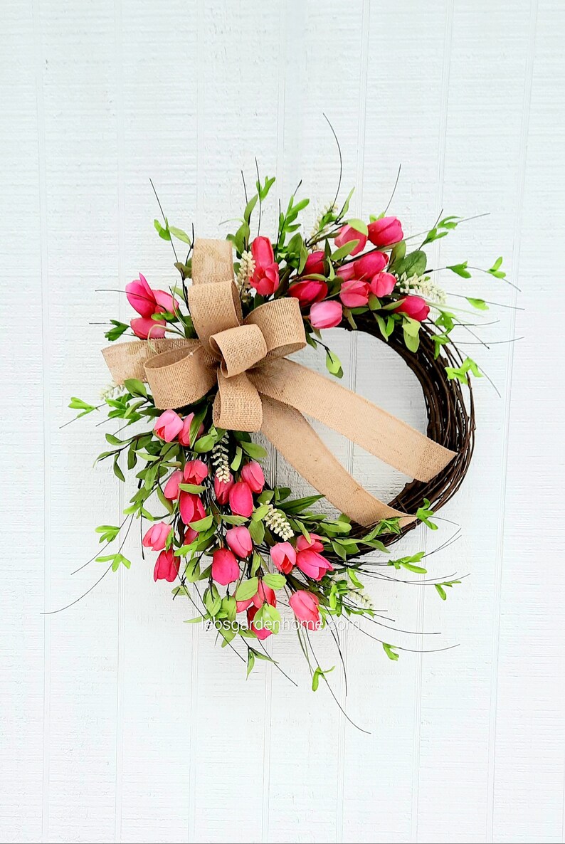 K&ouml;nnte beinhalten: Ein rosafarbener und gr&uuml;ner Blumenkranz mit einer Schleife aus Sackleinen. Der Kranz ist aus Weinreben gefertigt und mit rosa Tulpen und gr&uuml;nen Bl&auml;ttern verziert.