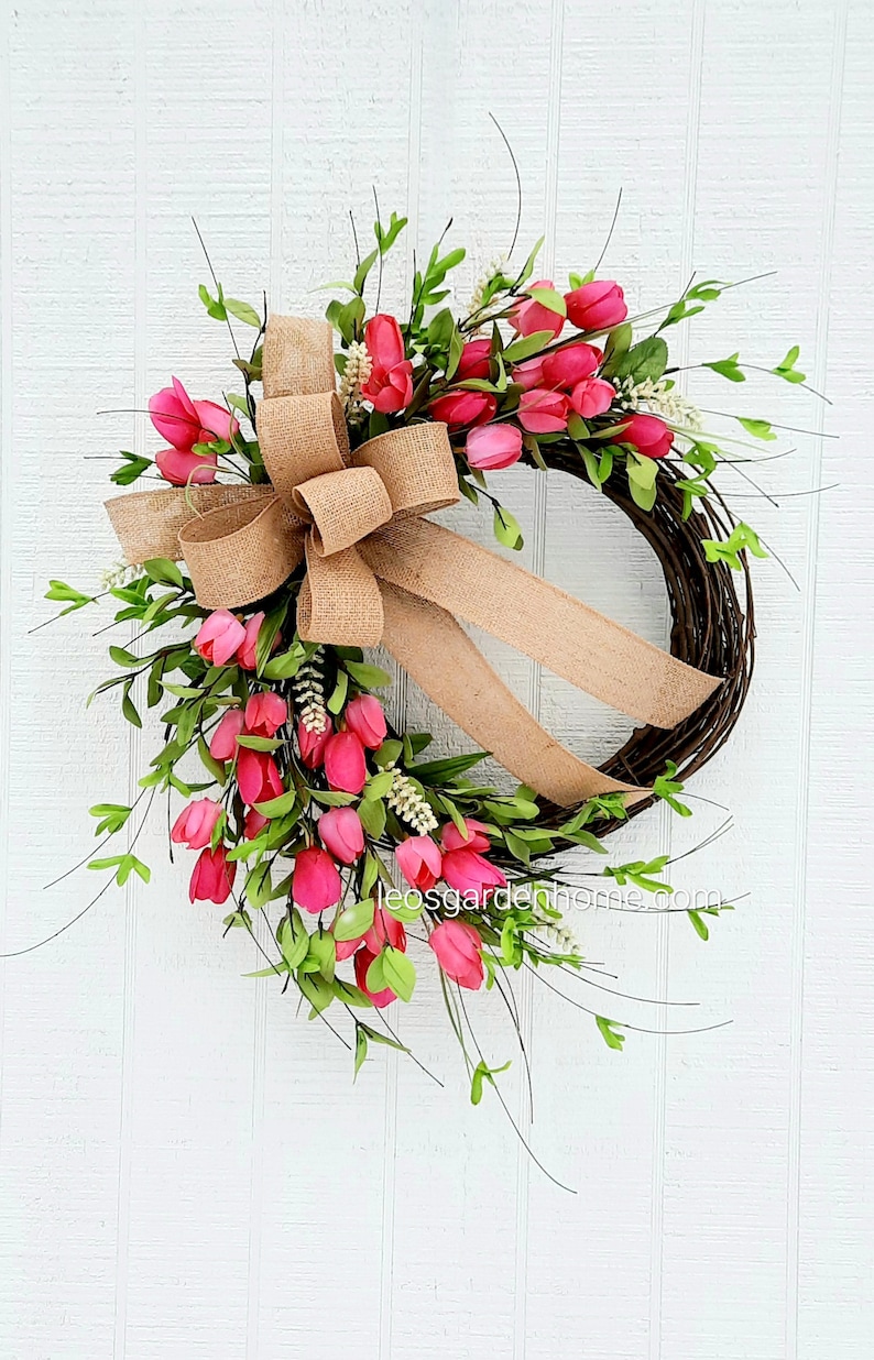 K&ouml;nnte beinhalten: Ein Fr&uuml;hlingskranz mit rosa Tulpen und Gr&uuml;n, verziert mit einer Schleife aus Sackleinen. Der Kranz ist auf einer Basis aus Weinreben gefertigt.