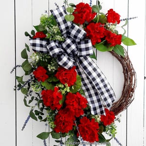 May include: A red geranium wreath with a black and white checkered ribbon bow. The wreath is made on a grapevine base and includes sprigs of lavender.