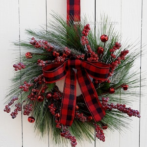 May include: A festive Christmas wreath with green pine needles, red berries, and small red bells. A large red and black buffalo plaid bow is centered on the wreath, with a matching ribbon for hanging. The wreath hangs against a white wooden background.