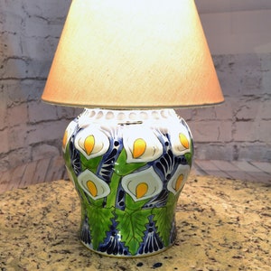 May include: A ceramic table lamp with a white fabric shade. The lamp base is decorated with a colorful floral pattern of white, yellow, green, and blue flowers.