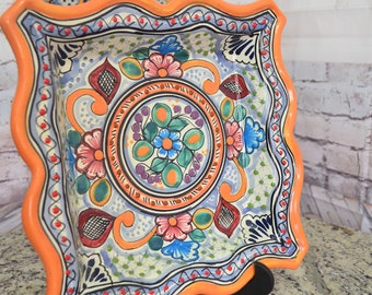 Puede incluir: Un plato cuadrado de cerámica con un borde festoneado. El plato está decorado con un colorido patrón floral en tonos azules, verdes, rosas y rojos. El plato está sobre un soporte negro.